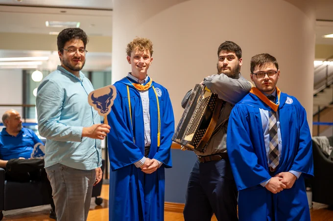 Four young men pose indoors; two in blue graduation gowns, one holding an accordion, and another with a buffalo fan. A seated man in blue is visible in the background.