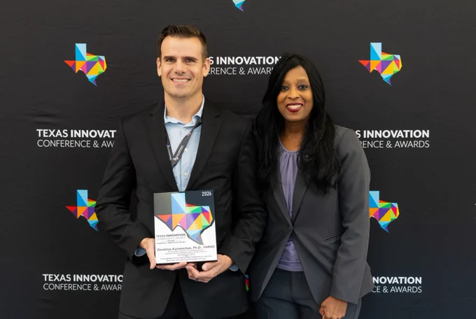 Two individuals are standing in front of a backdrop with "Texas Innovation Conference & Awards" written on it. One person holds a plaque. Both are wearing business attire.