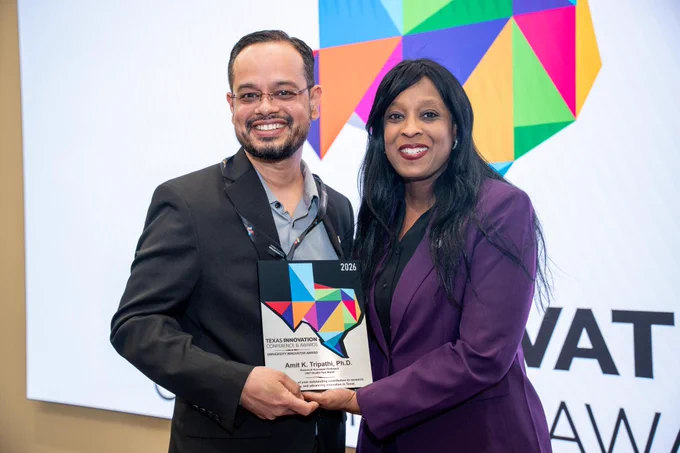 Two people are posing for a photo, smiling, and holding an award together. The background features a colorful, abstract geometric design.