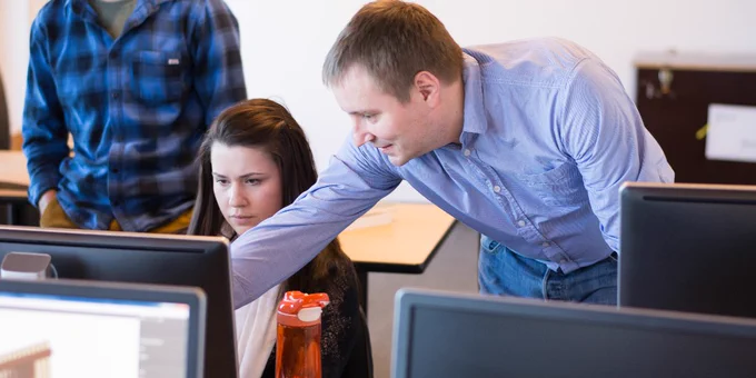 A person in a blue shirt is assisting another individual at a computer in a classroom setting. Other participants are in the background, and a water bottle is on the desk.