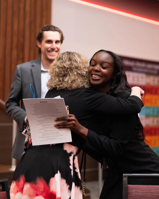 Two women warmly embrace in a congratulatory hug while holding a document, with a man standing nearby in a formal setting.