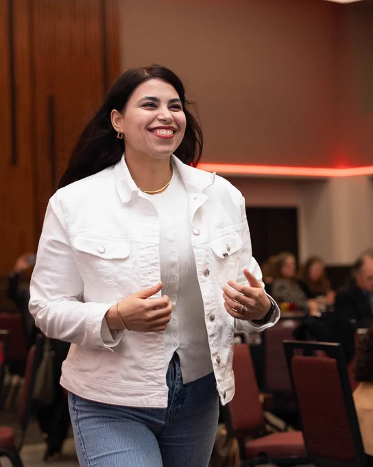 Person wearing a white denim jacket and blue jeans smiles brightly as she is walking towards the stage to accept her award.