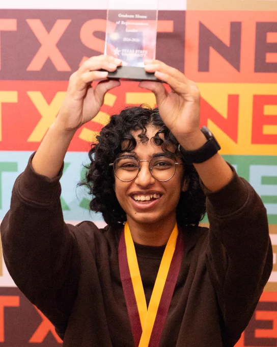 Student holding up a Graduate House of Representatives award triumphantly above her head in front of a colorful TXST backdrop. She smiles brightly