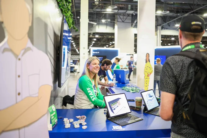 Staff member smiles at a trade show booth, showing tablet demos to a visitor across a blue counter in a busy expo hall.
