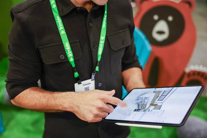 Person wearing a Trimble badge points at a tablet showing a 3D room design, with a cartoon bear display blurred in the background.