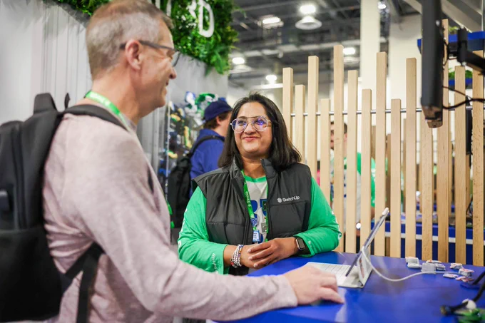 At a trade show booth, a woman in a SketchUp vest smiles while speaking with a man using a tablet at a blue counter.