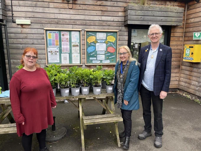 Outside a wooden building, three people stand around a wooden table in a small courtyard. The table holds many plants in pots. Among the three people is the outgoing Mayor of Trafford, Jane Brophy.
