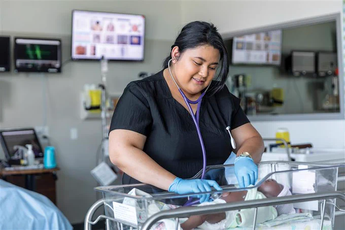 A smiling healthcare professional uses a stethoscope on a newborn in a hospital nursery. The room contains medical equipment and monitors in the background.