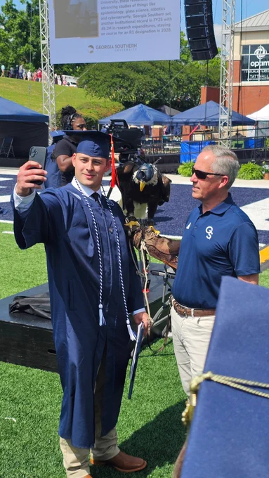 A male graduate takes a selfie with Freedom II, a live bald eagle mascot, at commencement.