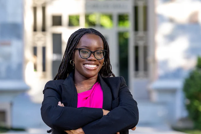 Emory Class Orator Joanna Louis-Ugbo smiles in a black jacket and pink shirt with Emory's pink marble buildings blurred behind her