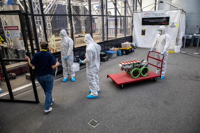 Inside a workspace at NASA's Kennedy Space Center, three people wearing full white protective suits, face shields, gloves, and blue shoe covers stand near a large enclosed clean room structure. One person in regular clothing opens a glass door leading into the area. Another person in protective gear pushes a red cart carrying a small, wheeled robotic device with green wheels and equipment mounted on top. Nearby are boxes, tools, and a white tent-like structure with warning and safety signs attached to it. Photo credit: Kim Shiflett