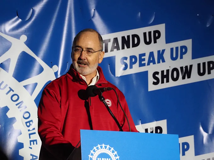 UAW president Shawn Fain in red jacket speaking at a podium with a backdrop reading 'Stand Up Speak Up Show Up'.