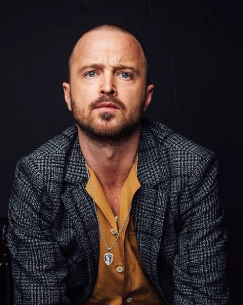 Professional headshot of Aaron Paul. He’s wearing an orange shirt and a grayish blazer with a silver necklace, looking directly at the camera.