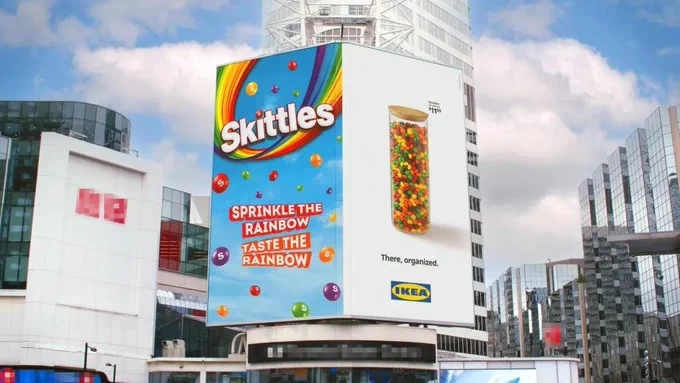 Large city billboard featuring colourful Skittles and IKEA advertisements on a modern skyscraper surrounded by reflective glass buildings under a cloudy blue sky.