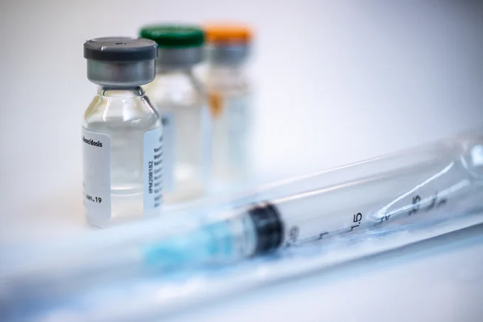 Close-up of a vaccine vial with a syringe in the foreground and additional vaccine vials blurred in the background.