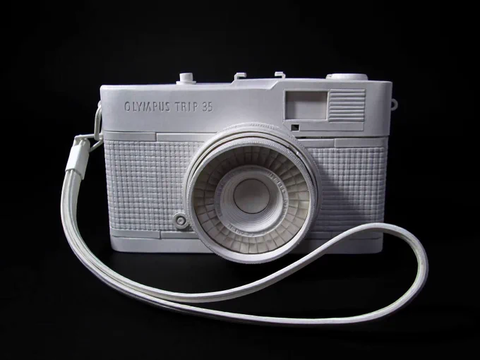 White paper sculpture of an Olympus Trip 35 camera model with a strap, displayed against a black background.