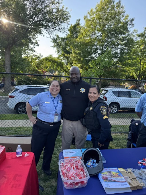 Three individuals in uniform pose outdoors at an event. They are standing behind a table with giveaways, including a basket of keychains. Two are wearing law enforcement uniforms, and one is wearing a security uniform. Trees and parked cars are visible in the background.