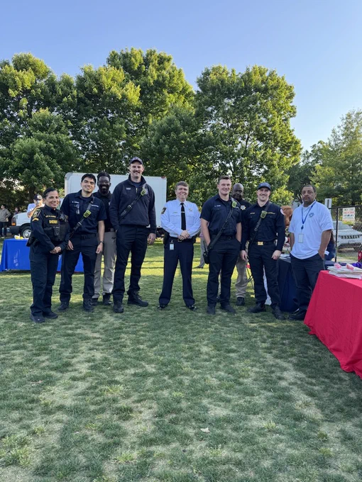 A group of uniformed police officers and firefighters standing on grass at an outdoor event, surrounded by display tables. Trees and a blue sky are visible in the background.
