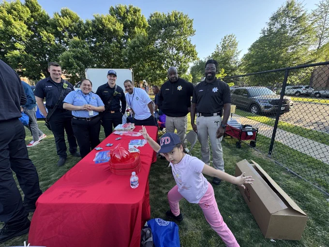 The image shows a group of people, including several in uniform, standing and smiling around a table covered with a red cloth. The table has a red fire helmet, brochures, and bottled water. A child is playfully stretching near the table. They are outdoors on a grassy area with trees and a fence in the background.