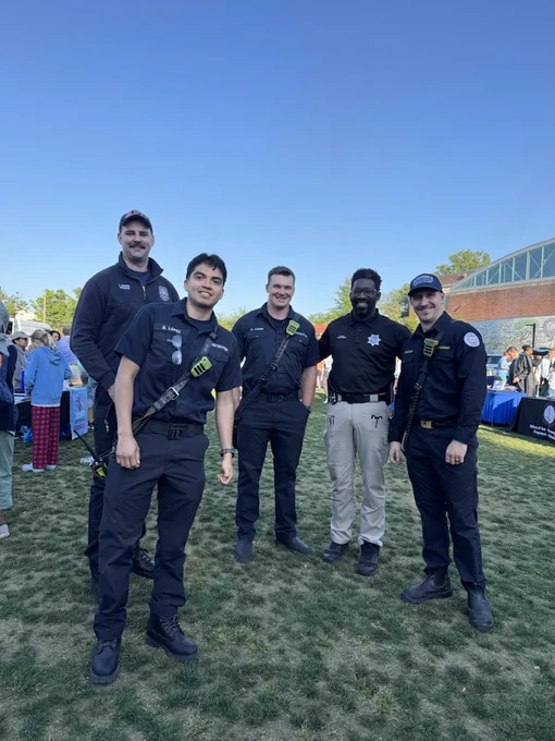 A group of five individuals in uniform stand on grass, smiling at an outdoor event. They are in front of tables with displays and people in the background.
