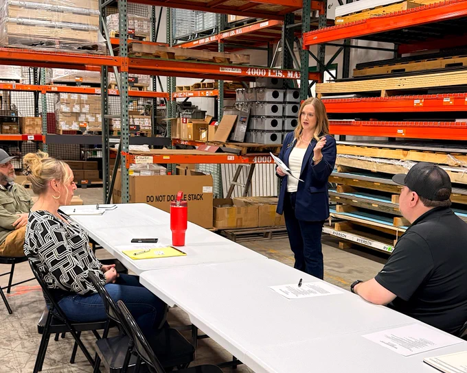 A person stands and speaks in a warehouse setting, addressing a group seated at a table. Shelves with various boxes and items are visible in the background.