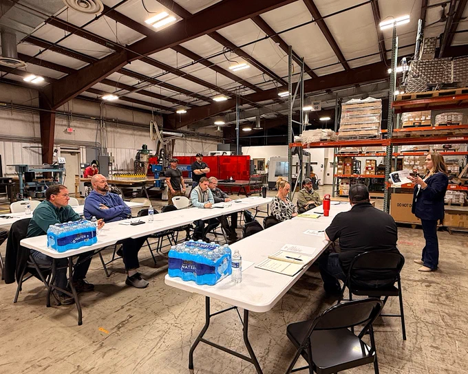 A group of people sitting around two white tables arranged in an L-shape inside a large industrial workshop. The space includes machinery and shelves stocked with supplies. A person at the front holds a paper, appearing to speak to the group. Two stacks of water bottles are on the tables.
