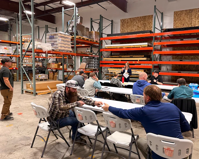 A group of people sit and stand in a large warehouse space with metal shelves stocked with various materials. They are gathered around several tables with white folding chairs. One person distributes papers to others seated at the tables. The setting has a casual, industrial feel.