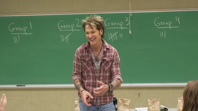 John Maag smiles while leading his class with a large green chalkboard and table with small brown paper bags on it behind him