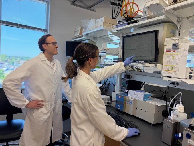 Two people in lab coats are in a laboratory. One is pointing at data displayed on a monitor, while the other observes. The room has various lab equipment.