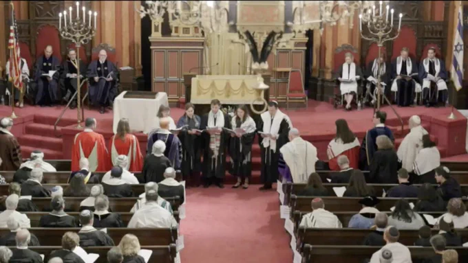 A formal ceremony takes place in a beautifully decorated synagogue, attended by a large congregation. Participants are dressed in ceremonial robes, suggesting a significant religious or community event.