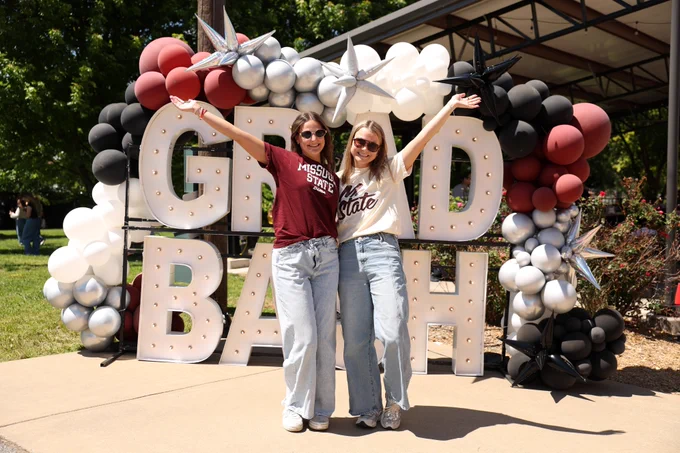 Two people stand smiling with arms raised in front of a large "GRAD BASH" sign decorated with balloons. Both are wearing "Missouri State" T-shirts. They are outdoors on a sunny day.