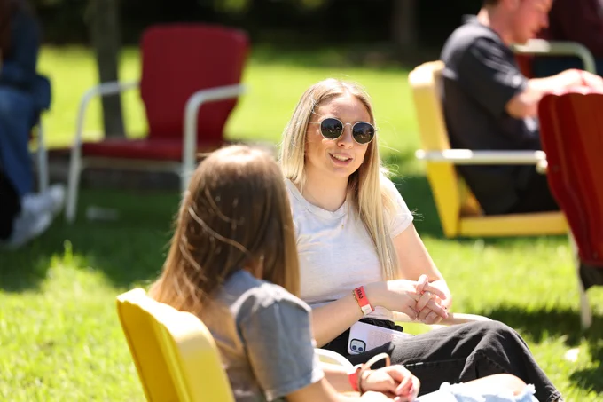 Two people are seated outdoors on sunlit chairs in a grassy area. One is wearing sunglasses and smiling toward the other person, both appear relaxed. Other individuals can be seen in the background, with blurred trees and more seating.