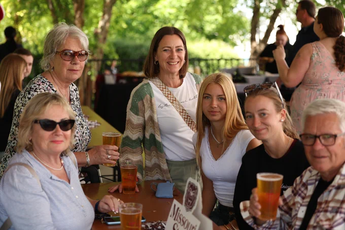 Six people, mainly women, at an outdoor gathering. A smiling woman in a striped cardigan, a blonde woman, and another woman hold amber drinks on a brown table against a green background.
