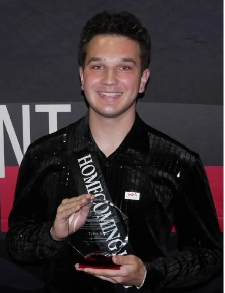 Tyler Rose smiles while holding a transparent award plaque. He is wearing a dark shirt and a sash that reads "Homecoming.