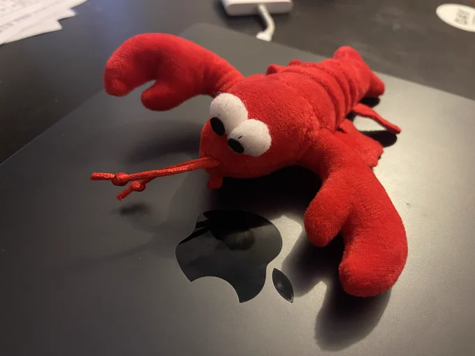 Close-up photo of a small red plush lobster toy resting on the lid of a dark-colored Apple MacBook. The lobster has oversized white cartoon eyes with black pupils, large soft claws, and a red string extending from its mouth like antennae or whiskers. The plush fabric appears fuzzy and slightly worn, giving it a playful, well-used look.

The reflective black Apple logo is visible beneath the toy near the center of the laptop. The scene is lit with warm indoor lighting, creating soft shadows and emphasizing the bright red color of the plush against the matte dark surface of the MacBook. The background is slightly out of focus, with parts of a desk and cables faintly visible.