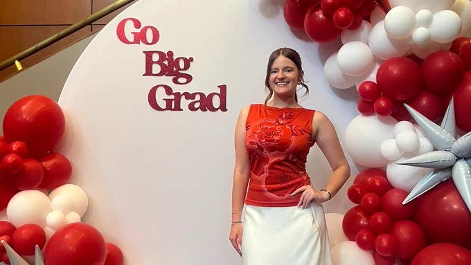 Woman stands in front of white banner and red and white balloons, tet says Go Big Red.