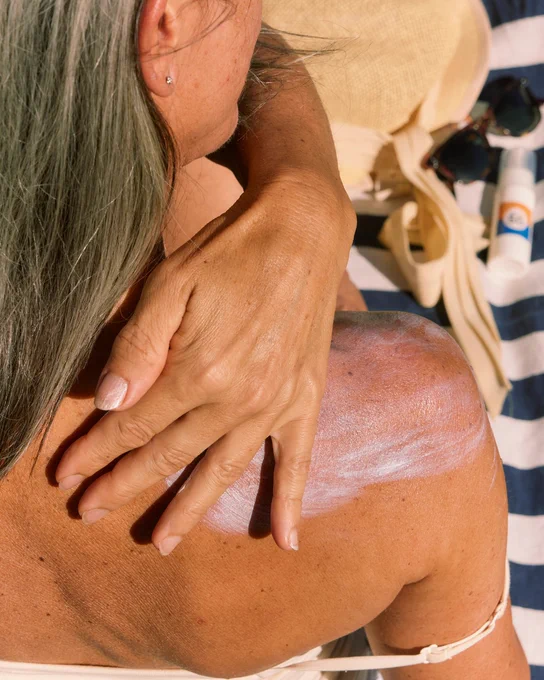 A person applies sunscreen to their shoulder while sitting on a beach towel with a striped pattern. Nearby are sunglasses, a hat, and a sunscreen bottle.