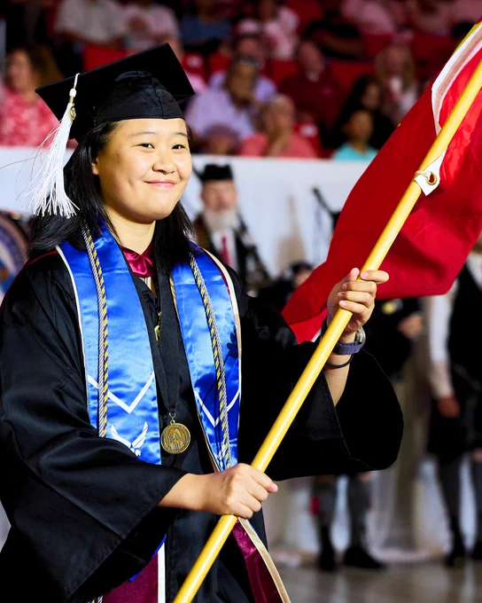 A graduate in a cap and gown, adorned with blue and gold stoles and a medal, holds a flag during commencement ceremony. A crowd watches in the background.