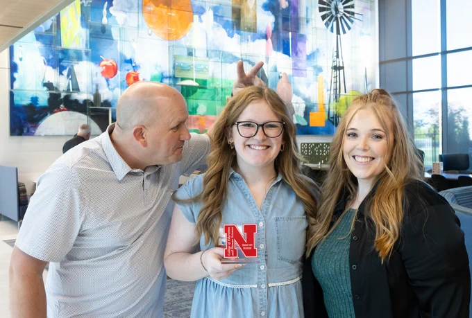 Paige Wooster’s dad gives her bunny ears  and looks at her while she holds her Graduate with Distinction award and smiles for a photo with her mom on the other side of her in the Living Room area of Carolyn Pope Edwards Hall