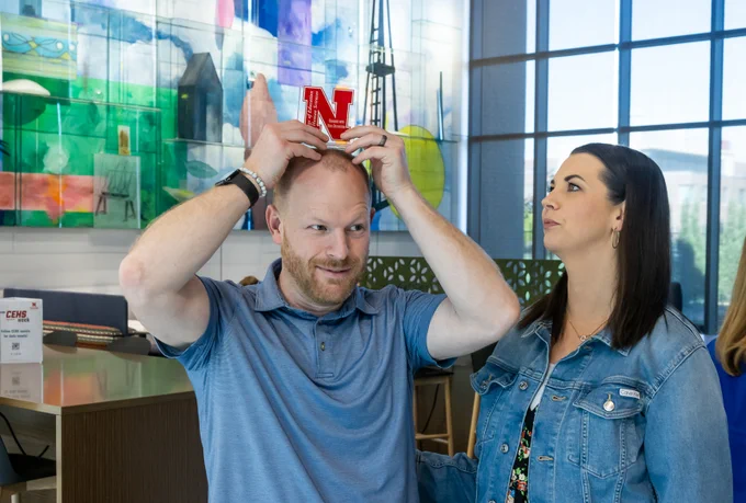 Eric Jacob attempts to balance his Graduate with High Distinction award on his head as his wife watches while preparing to take a photo together in the Living Room area of Carolyn Pope Edwards Hall