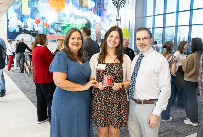 A student holds her Graduate with Distinction award while posing for a photo with her parents near the Living Room area of Carolyn Pope Edwards Hall which is full of other students and their families