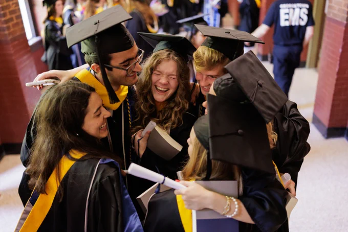 A group of six master's students in graduation regalia huddle into a hug.