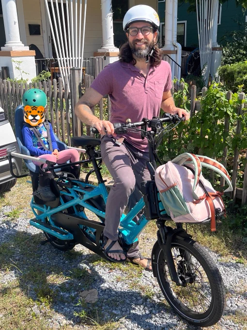 A person wearing a helmet rides a cargo bike with a child seated in the back. The child is wearing a helmet and sunglasses. There is a bag attached to the front of the bike. They are on a gravel path in front of a house with a small fence.