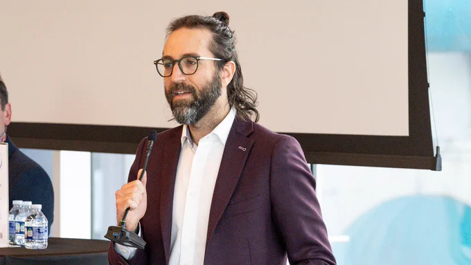 A person with long hair tied back stands in a room, holding a microphone. They are wearing glasses and a dark jacket over a white shirt. Bottles of water are visible on a table beside them.