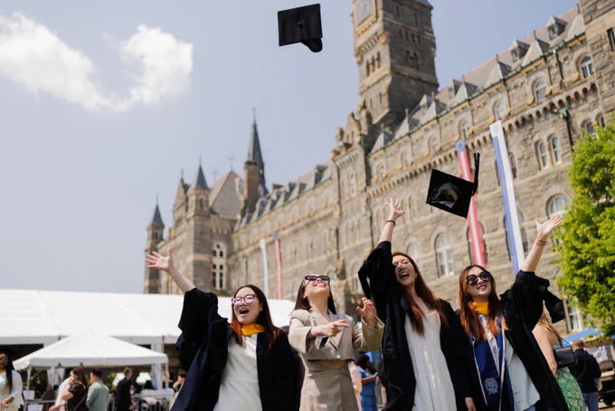 Master's students wearing graduation regalia throw graduation caps into the air in front of Healy Hall.