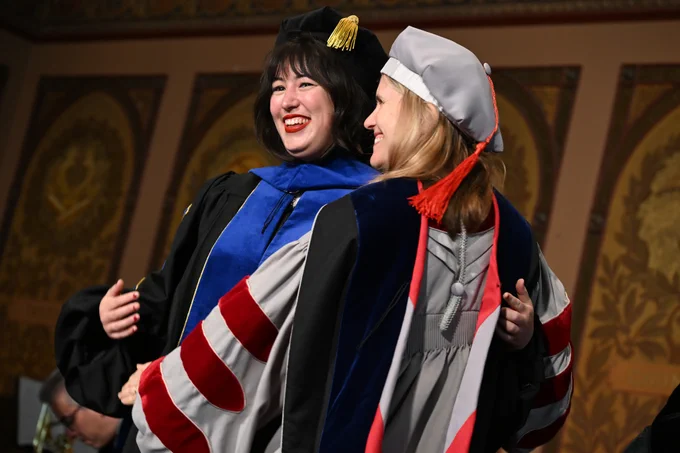 A Ph.D. graduate smiles and side-hugs her doctoral advisor. Both are wearing a doctoral tam, hood and robe.