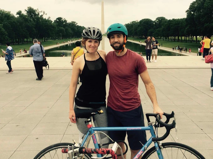 Two individuals wearing helmets stand together with a bicycle in front of the Washington Monument. The Reflecting Pool and several people are visible in the background.