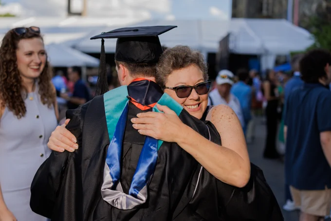 A graduating master's student, seen from behind, hugs a loved one outside the graduation tent.
