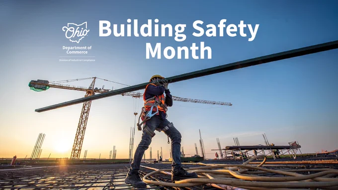 A construction worker stands on a building site at sunset, holding a large metal beam. Several cranes are visible in the background. The text reads "Building Safety Month" and includes the logo for the Ohio Department of Commerce, Division of Industrial Compliance.