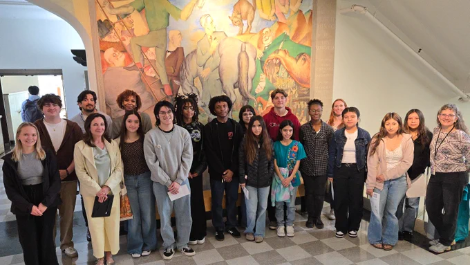 20 students stand together and pose for a photo in front of a large mural.